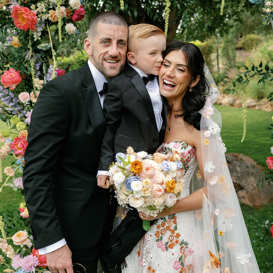 A happy couple and child pose outside with The Promise florals by Eos Blooms in the background.