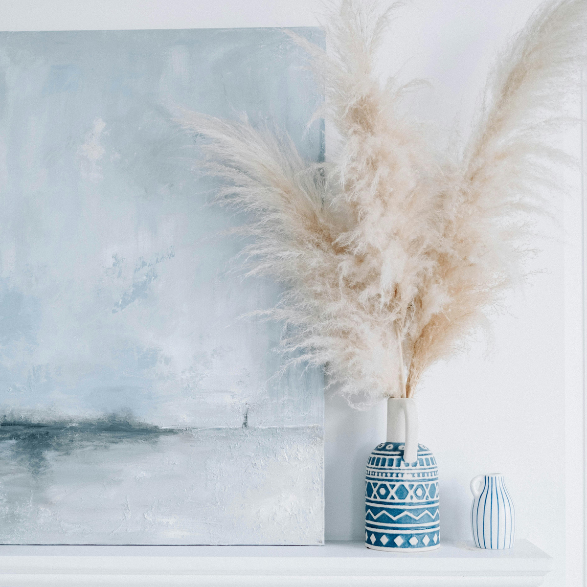 A blue abstract painting and pampas grass in patterned vases on a white shelf.