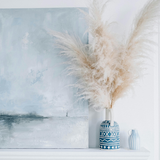A blue abstract painting and pampas grass in patterned vases on a white shelf.