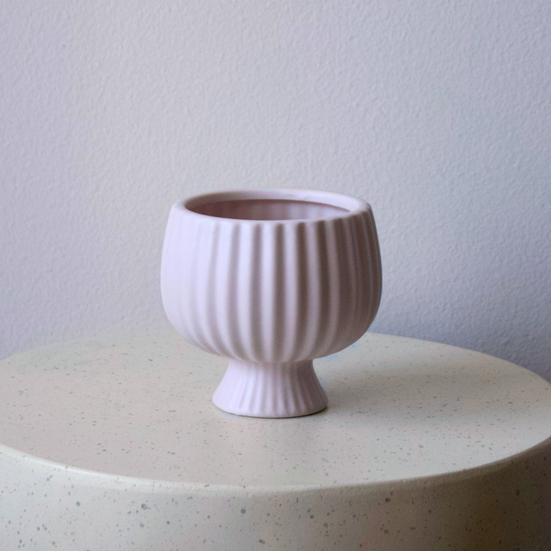 The Eos Blooms Small Blush Pedestal rests on a speckled surface beside a pale wall and vase.