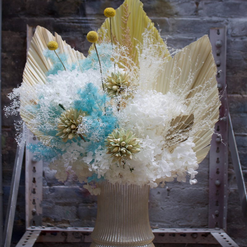 The Calypso by Eos Blooms: preserved flowers with palm leaves and blue in a ribbed vase.