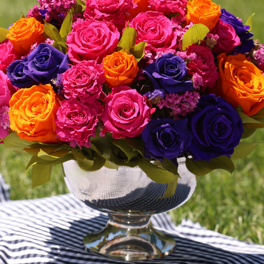 The Mariposa by Eos Blooms holds vibrant preserved flowers on striped cloth outdoors.