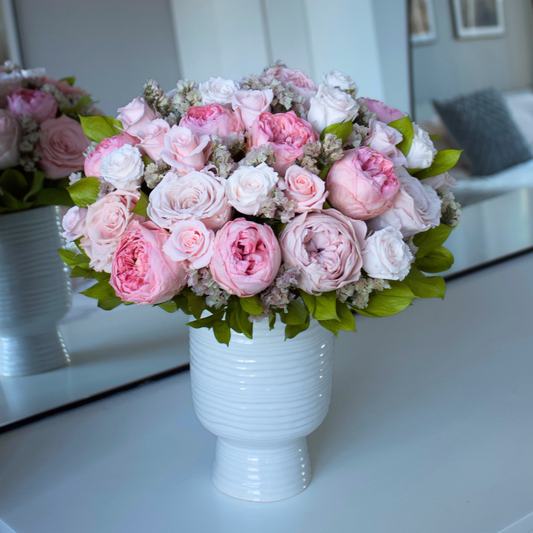 The Paloma by Eos Blooms, a white vase with preserved roses and greenery, sits on a table.