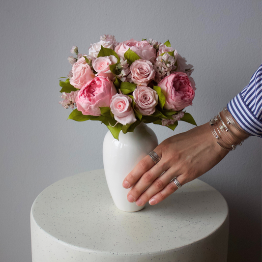 A hand adjusts The Paloma (Petite) by Eos Blooms on a round, light-colored table.