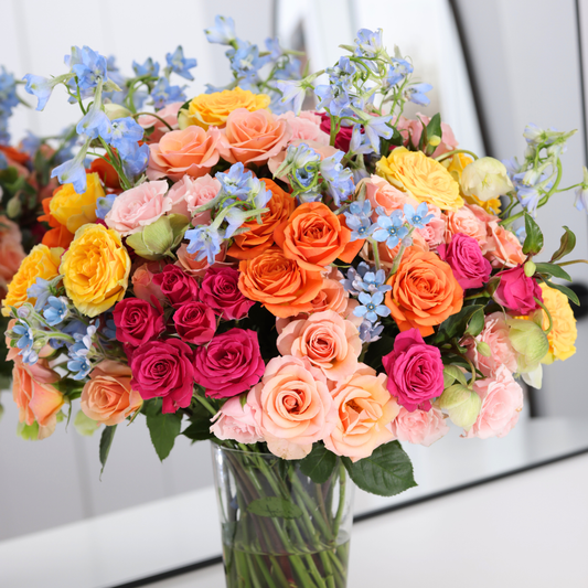 A bouquet of colorful roses and blue flowers in a glass vase on a white surface.