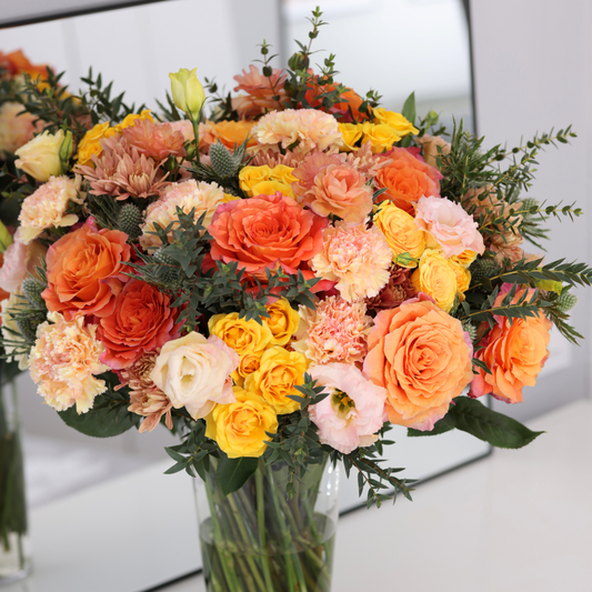 A bouquet of orange, yellow, and peach roses and carnations in a glass vase.