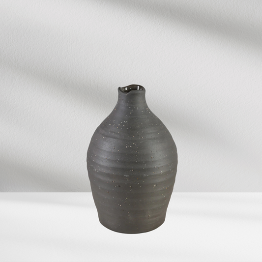 A black Eos Blooms Tikal Vase with a small spout sits on a white surface against a light background.