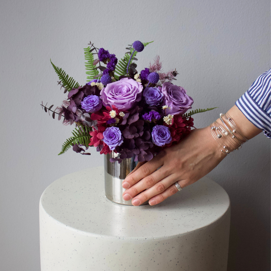 A hand arranges the Eos Blooms Ultraviolet (Petite) on a round, white table.