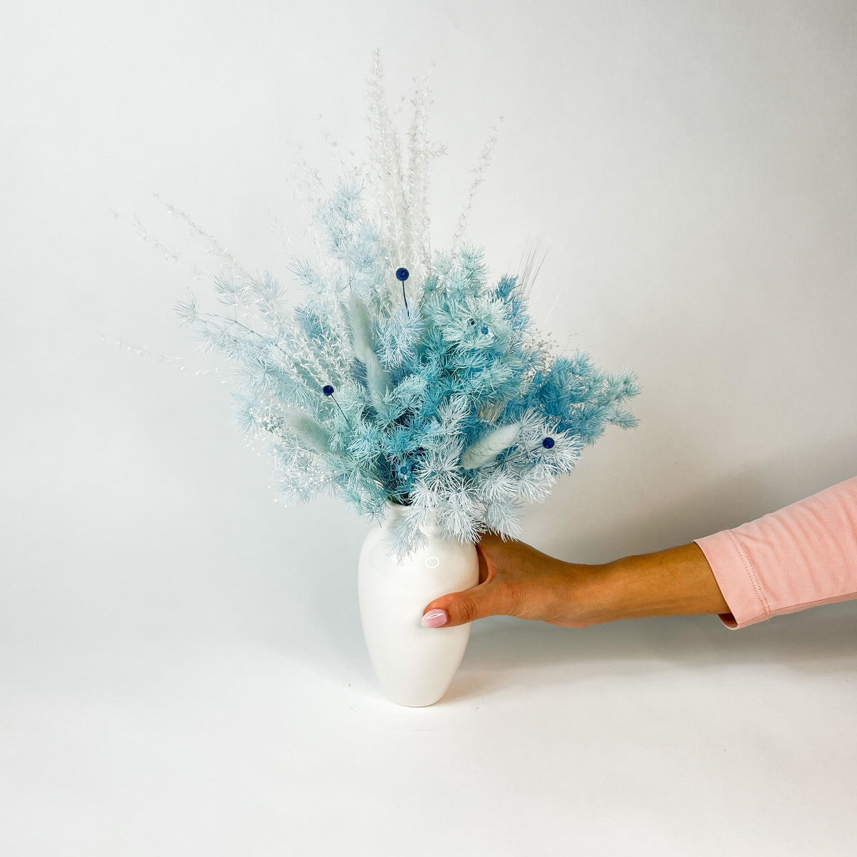 A hand holds Eos Blooms' Serenity, a white vase with Forever Flowers, against a white background.